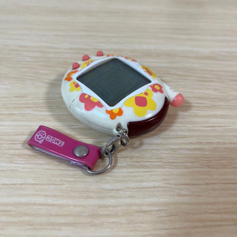 May include: A vintage handheld digital pet game with a white body, adorned with yellow and pink floral patterns. It features a small screen and a pink strap with the text "TMGC". The device has a red and brown base.