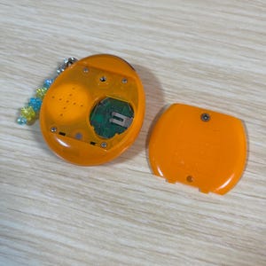 May include: An open, orange handheld electronic device with a detached cover. The device has a screen, buttons, and internal components visible. A beaded keychain is attached to the device. The device is on a light wood-grain surface.