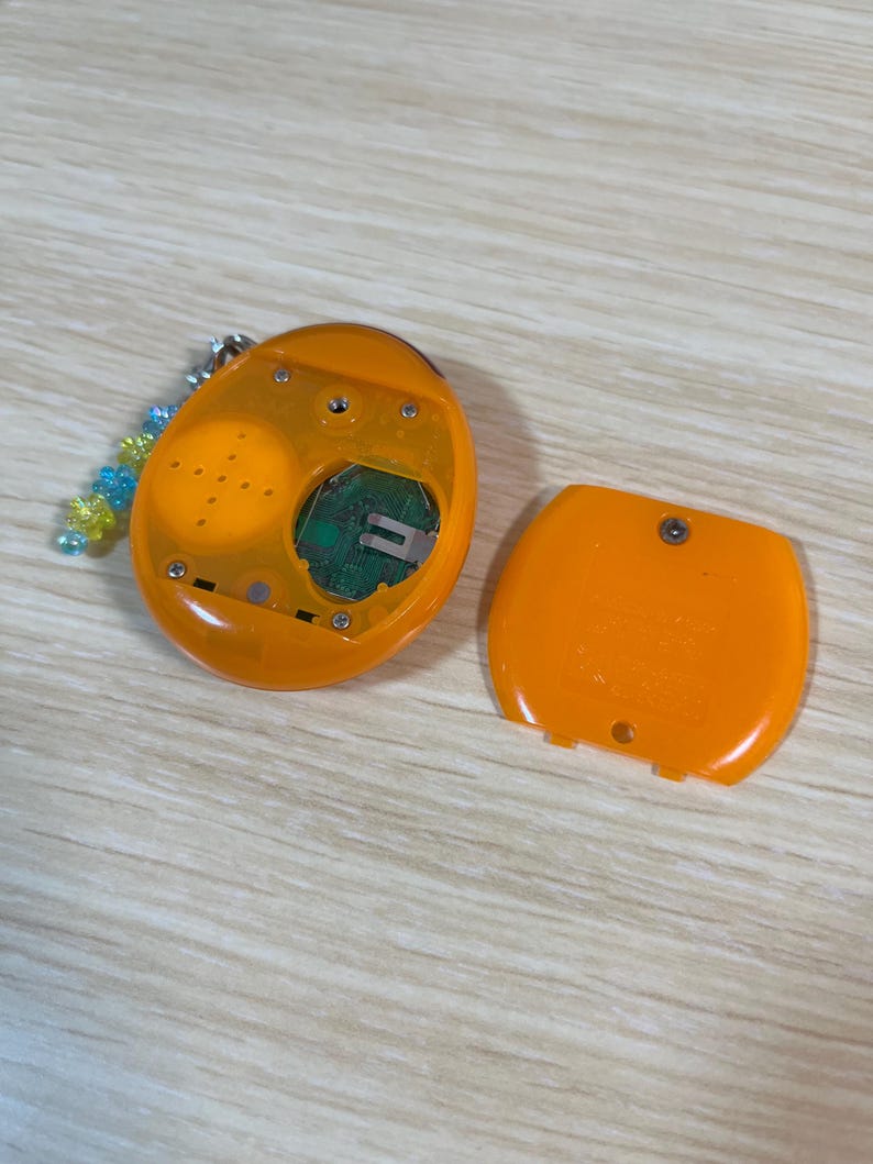 May include: An open, orange handheld electronic device with a detached cover. The device has a screen, buttons, and internal components visible. A beaded keychain is attached to the device. The device is on a light wood-grain surface.