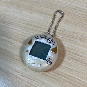 May include: A pearlescent white Angel Gotcha virtual pet toy with a silver chain. The egg-shaped device features a small screen displaying a star, gold wing accents, and the text "ANGEL GOTCHA". Three buttons are below the screen.