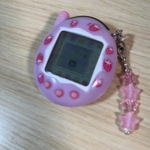 May include: A light purple handheld digital pet game with a small screen and pink strawberry accents. The device has three pink buttons and a silver chain with pink star-shaped charms attached. The screen displays a pixelated graphic.