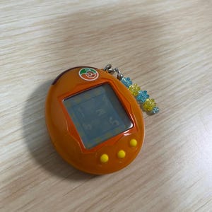 May include: Orange handheld digital pet game with a small screen displaying numbers and letters. The device has three yellow buttons and a keychain with blue and yellow beads. The top of the device has a small orange logo.