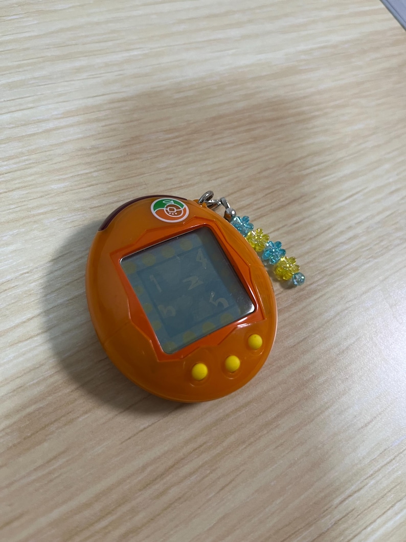 May include: Orange handheld digital pet game with a small screen displaying numbers and letters. The device has three yellow buttons and a keychain with blue and yellow beads. The top of the device has a small orange logo.