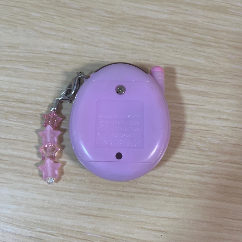 May include: A light purple handheld electronic game with a pink antenna and a decorative star charm. The device has a small screen and buttons, and is likely a vintage or retro toy. The charm features pink star-shaped beads.