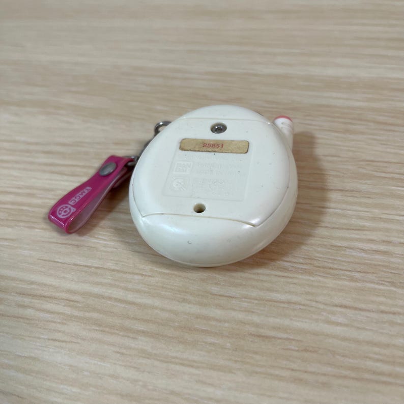 May include: A white, egg-shaped handheld electronic game with a pink keychain. The game has a small screen and several buttons. The keychain is pink with the word "Tamagotchi" printed on it. The game is on a light wood surface.