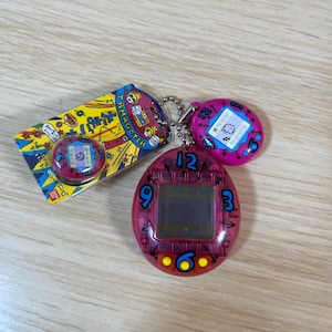 以下が含まれることがあります： デジタル画面を備えたピンク色のたまごっちバーチャルペットおもちゃ。3、6、9、12の数字で囲まれています。小さなピンク色のたまごっちが付属しており、日本語のテキストが書かれたパッケージ版もあります。