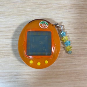 May include: Orange handheld digital pet game with a small screen and three yellow buttons. The device has a silver chain with blue and yellow flower-shaped beads attached. The screen displays numbers.
