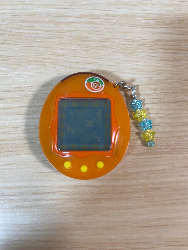 May include: Orange handheld digital pet game with a small screen and three yellow buttons. The device has a silver chain with blue and yellow flower-shaped beads attached. The screen displays numbers.