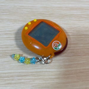 May include: An orange handheld digital pet game with a small screen and yellow buttons. It has a decorative charm with blue and yellow flower-shaped beads attached to a silver clasp. The device is on a light wood surface.