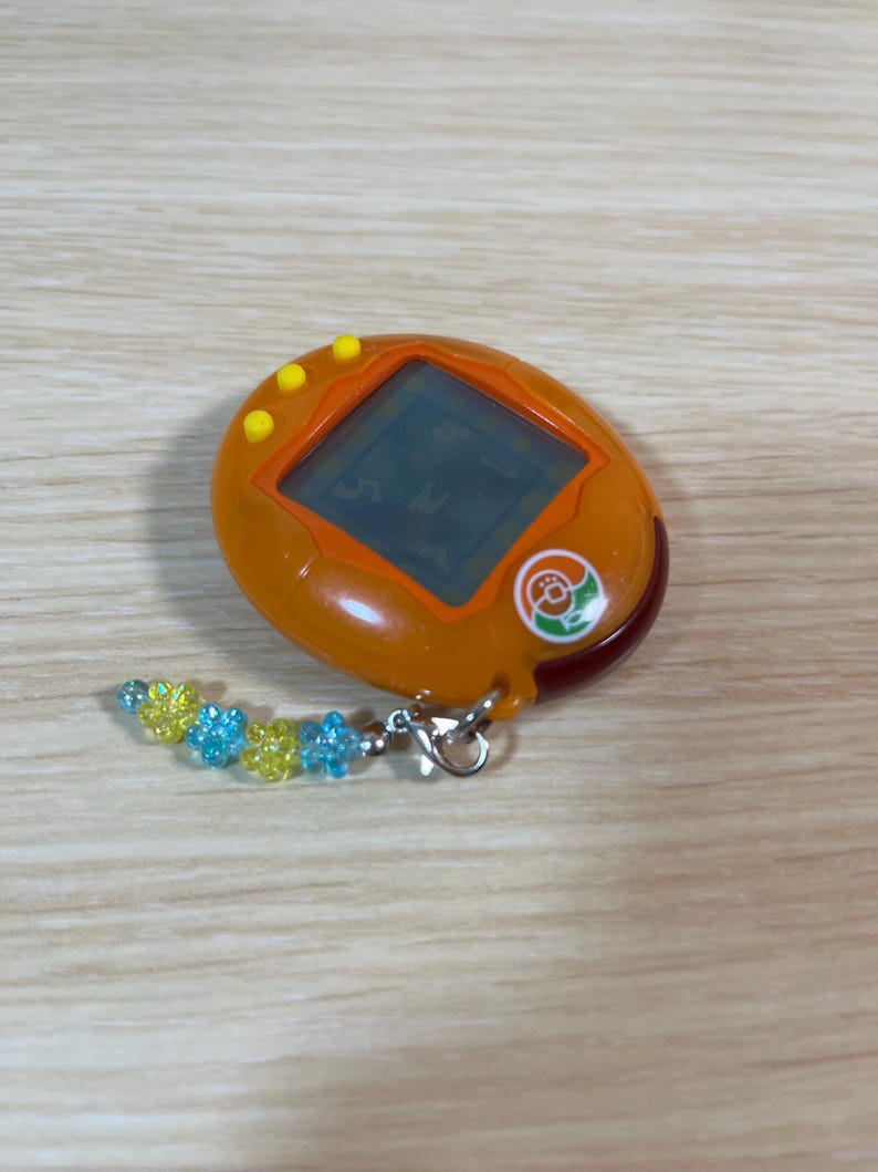 May include: An orange handheld digital pet game with a small screen and yellow buttons. It has a decorative charm with blue and yellow flower-shaped beads attached to a silver clasp. The device is on a light wood surface.