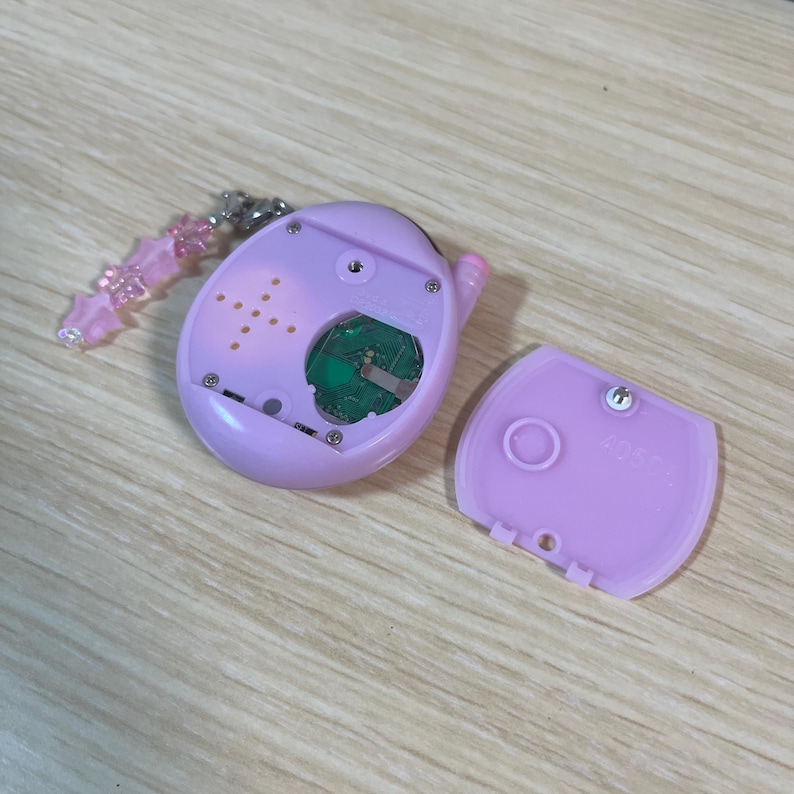 May include: A disassembled, light purple electronic device with a green circuit board visible. The device has a detached cover with the number "40504" printed on it. A pink star-shaped charm is attached.