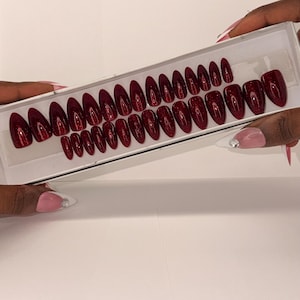 May include: A set of almond-shaped press-on nails in a deep, dark red colour with a subtle glitter finish. The nails are arranged in a white rectangular display box, featuring various sizes to accommodate different nail beds for a polished look.