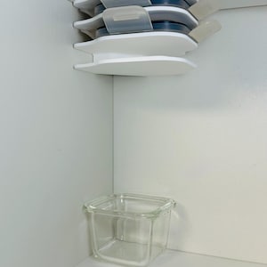 May include: A stack of white, tiered storage containers with clear lids and a clear glass square container. The containers are in a white cabinet corner. The lids are a light grey colour.