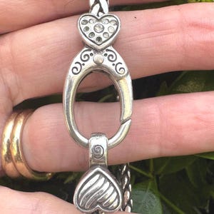 Pu&ograve; includere: Bracciale color argento con design a catena intrecciata. Presenta dettagli a forma di cuore, uno impreziosito da cristalli trasparenti. La chiusura &egrave; ovale con volute decorative.