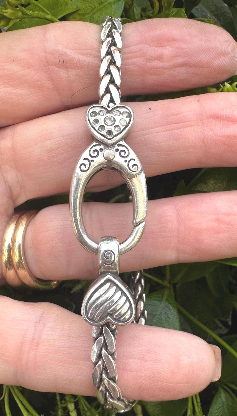 Pu&ograve; includere: Bracciale color argento con design a catena intrecciata. Presenta dettagli a forma di cuore, uno impreziosito da cristalli trasparenti. La chiusura &egrave; ovale con volute decorative.