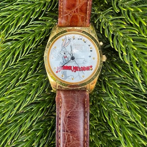 May include: Gold-tone Armitron watch with a brown leather band. The watch face features Bugs Bunny from Looney Tunes with the words "Merrie Melodies". The watch is set against a backdrop of green foliage.