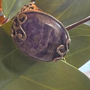 May include: Oval-shaped purple gemstone pendant with ornate silver-colored metalwork. The pendant is set against a backdrop of green leaves, highlighting the gemstone's color and texture. A unique piece of jewelry.