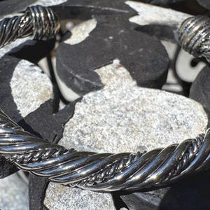 May include: A silver-toned, twisted metal cuff bracelet. The bracelet has a rope-like design with a polished finish. The ends of the bracelet have a thicker, ridged design. The bracelet is displayed on a background of gray and black stones.