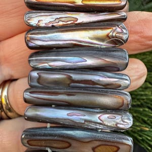 May include: A stacked bracelet composed of rectangular, iridescent abalone shell beads. The beads display a range of colors, including brown, gold, and blue, with a glossy finish. The bracelet is a piece of jewelry.