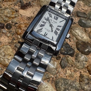 May include: A silver ACTIVA wristwatch with a rectangular face and a silver band. The watch face is white with black Roman numerals and hands. The watch is water resistant to 30M.
