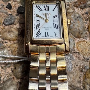 May include: Gold-toned Activa wristwatch with a rectangular face and a white dial featuring Roman numerals and hands. The watch has a water resist rating of 30M and a gold-toned band. The watch is displayed on a stone surface.