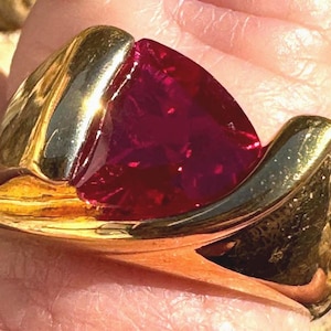 May include: A gold ring featuring a large, triangular-cut, deep red gemstone set centrally within a polished, smooth, yellow gold band that curves around the stone, creating a sleek, continuous metal frame around the vibrant red center.