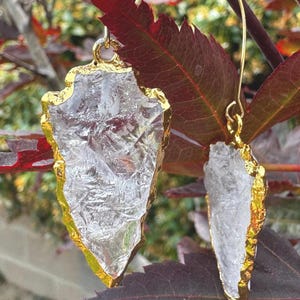 May include: A pair of gold-plated quartz earrings. The earrings are shaped like arrowheads with a clear, textured quartz center and a gold-colored border. The earrings hang from gold-colored ear wires, set against a backdrop of red and green leaves.