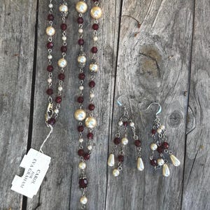 Puede incluir: Un conjunto de collar y pendientes con perlas de imitación color crema, cuentas de color burdeos y detalles metálicos plateados. El collar tiene un diseño de múltiples hilos con un colgante de borla. Los pendientes son colgantes.