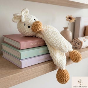 May include: A handmade, cream-colored crochet lamb toy with tan accents, resting on a stack of pastel-colored books. The lamb has a round head, floppy ears, and a long, textured body. The books are titled "The Little Bear's Journal", "Lullaby Land", and "Lavegood Funny Baby".