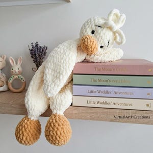 May include: A handmade, plush duck toy in cream and tan. The duck is lying on a stack of books with titles like "The Moon's Cradle" and "Little Waddles' Adventures." The toy has a textured, fluffy appearance, with a tan beak and feet. The overall theme is children's toys.