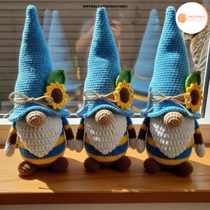May include: Three handmade crochet gnomes with blue hats, white beards, and sunflower accents. The gnomes have brown and yellow striped bodies and are displayed on a wooden surface. The text "VIRTUALARTHCREATIONS" is visible.