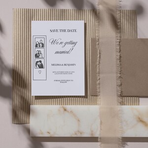 May include: A wedding invitation suite featuring a white card with the text "Save the Date" and "We're getting married!" with photo booth style images. The suite includes a brown envelope, ribbon, and a marble accent.