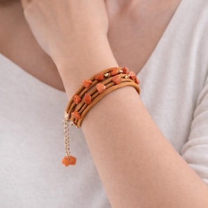 Könnte beinhalten: Mehrreihiges Wickelarmband aus hellbraunem Leder mit orange-roten Steinperlen und goldenen Akzenten. Das Armband hat einen kleinen Anhänger, der an einer goldenen Kette hängt.