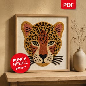 Pode incluir: Um padrão de punch needle em PDF apresentando uma cabeça de leopardo estilizada com manchas amarelas, laranja e pretas sobre um fundo de tecido creme. O design inclui olhos verdes, nariz preto e bigodes pretos, em uma moldura quadrada de madeira clara.