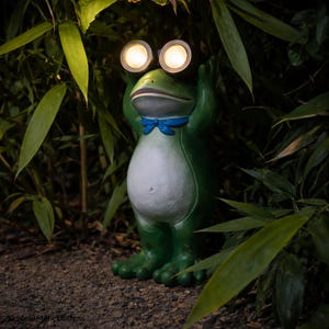 May include: A whimsical green frog statue with two round, illuminated lights for eyes, set amidst lush greenery. The frog has a white belly and a blue bow tie. The statue is designed for outdoor garden decor.