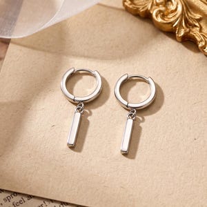 May include: A pair of silver-toned hoop earrings, each featuring a small, smooth, rectangular bar charm suspended from the bottom. The earrings have a polished metallic finish and a hinged closure mechanism, presenting a clean and simple design.