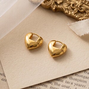 Puede incluir: Un par de pendientes de aro pequeños en tono dorado con forma de corazón. Los pendientes presentan una superficie lisa y reflectante con un mecanismo de cierre de bisagra, diseñados para ajustarse al lóbulo con una silueta redondeada.