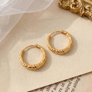May include: A pair of small gold hoop earrings with a textured, hammered surface. The circular earrings feature a hinged closure mechanism and a polished metallic finish, reflecting light across their uneven, dimpled exterior surface.