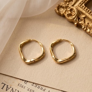 May include: A pair of gold-toned hoop earrings with a distinct square-like, irregular shape. The earrings feature a smooth, polished metallic surface with a continuous loop design and a hinged closure mechanism, showcasing a reflective finish.