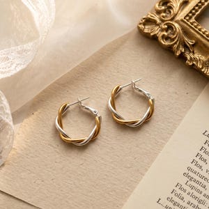 May include: A pair of hoop earrings featuring a twisted design with alternating silver and gold-toned metal strands. Each earring has a post-back closure mechanism and a circular shape, showcasing a two-tone metallic finish throughout the piece.