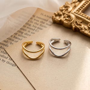 Op de afbeelding: Twee open ringen, één in een gepolijste goudtint en één in een gepolijste zilverkleur. Elke ring heeft een ontwerp met dubbele band en een gladde, afgeronde afwerking. De ringen liggen naast elkaar op een vel papier met gedrukte tekst.