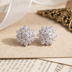 May include: A pair of silver-toned stud earrings featuring a floral design. Each earring is composed of multiple marquise-cut and round-cut clear white stones arranged in a layered, sunburst pattern that radiates from a central round stone.