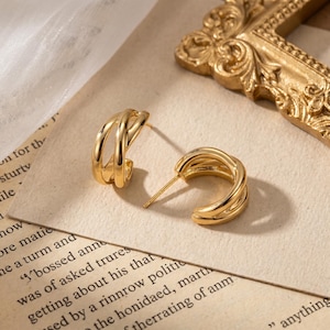 May include: A pair of gold-toned hoop earrings featuring a multi-layered, intertwined design. Each earring consists of three thin, curved bands that meet at the post, creating a dimensional, open-loop structure with a polished, reflective finish.