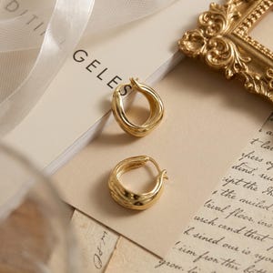 Könnte beinhalten: Zwei goldfarbene Creolen. Ein Ohrring hat ein verdrehtes Design, der andere eine geriffelte Textur. Die Ohrringe werden auf einer cremefarbenen Oberfläche mit schwachem Text und einem goldfarbenen dekorativen Rahmen präsentiert.