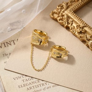 May include: A gold-toned double ear cuff connected by a delicate chain. Each cuff features an irregular, organic surface texture and is embellished with small, four-pointed star motifs, each set with a tiny, clear, sparkling stone in the center.