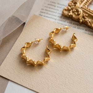 Może przedstawiać: Para złotych kolczyków w kształcie kół z charakterystycznym falistym, karbowanym brzegiem. Kolczyki są okrągłe z przerwą i posiadają przezroczyste plastikowe zatyczki. Powierzchnia metalu jest gładka i ma wykończenie o wysokim połysku.