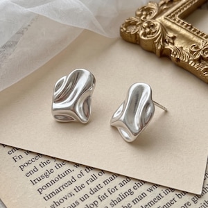 May include: A pair of silver-toned stud earrings featuring an irregular, molten-like organic shape with a smooth, reflective surface. The earrings have a post-back closure and are displayed on a piece of paper with printed text.