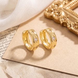Puede incluir: Un par de pendientes de aro dorados con acabado pulido y motivos de estrellas grabados. Cada estrella tiene en el centro una pequeña piedra preciosa transparente de talla redonda. El interior de cada aro lleva grabado el sello 925 de plata de ley.