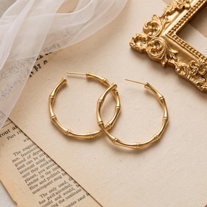 May include: A pair of gold-toned hoop earrings featuring a textured bamboo-inspired design. The earrings are circular with a polished metallic finish, showcasing segmented nodes that mimic the appearance of natural bamboo stalks throughout the hoops.