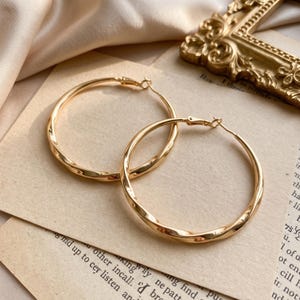 Puede incluir: Un par de pendientes de aro dorados con un diseño de metal trenzado. Los pendientes tienen forma circular con una superficie pulida y reflectante, y cuentan con un cierre de poste estándar, mostrando una textura en espiral continua.